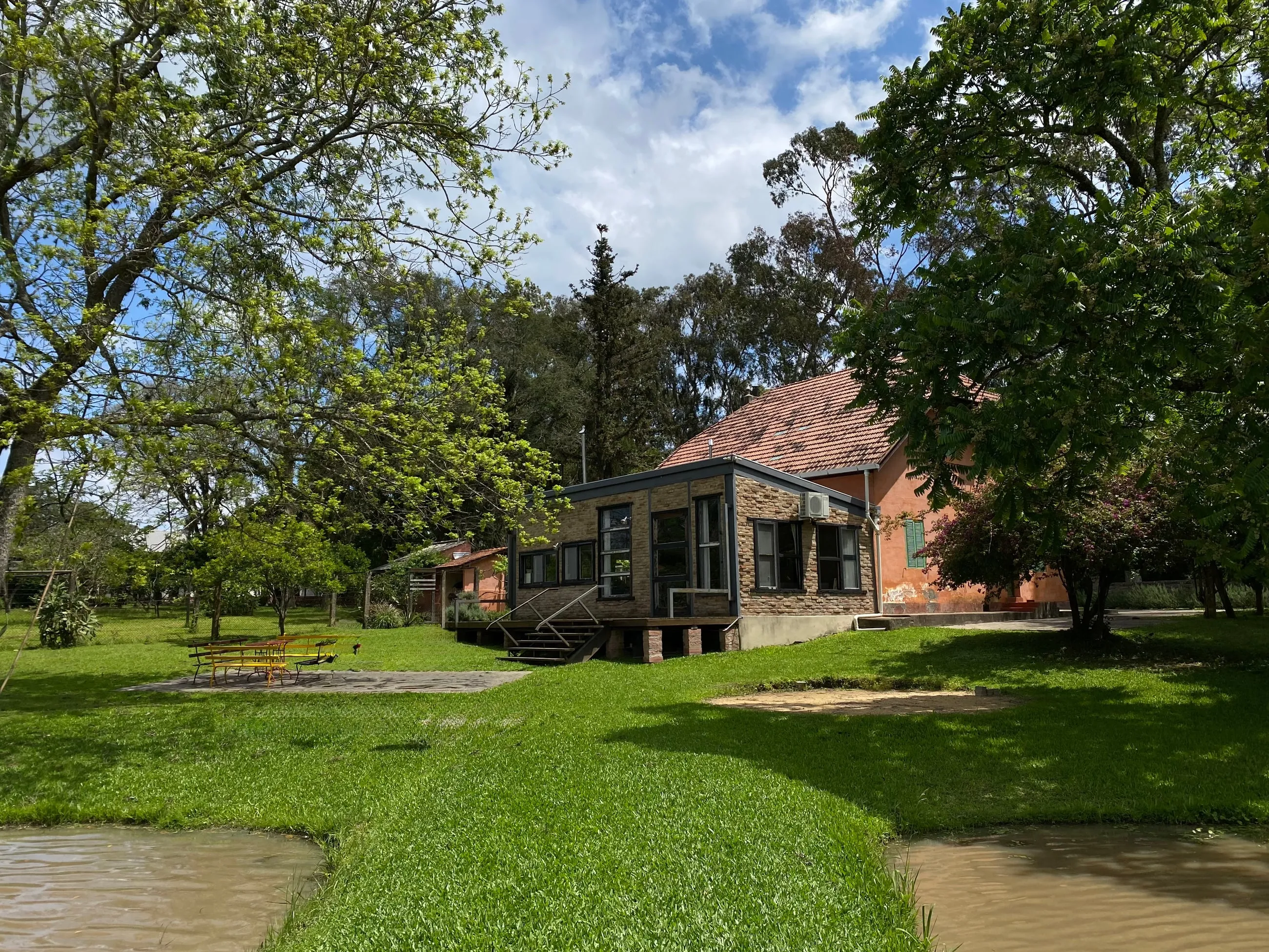A Casa Micael na Granja Nenê, entre o gramado, as árvores e a água, sob o céu azul.